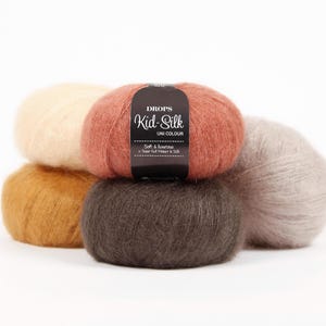 Puede incluir: Cinco ovillos de lana esponjosa en tonos crema, óxido, amarillo mostaza, marrón oscuro y gris claro. Una etiqueta negra en el ovillo central de color óxido dice DROPS Kid-Silk UNI COLOUR Soft & luxurious Super Kid Mohair & Silk.
