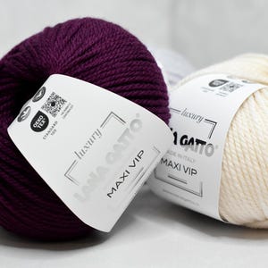 Può includere: Due gomitoli di filato Lana Gatto Maxi VIP, uno viola scuro e uno color crema. Ogni gomitolo presenta un'etichetta di carta bianca con il testo 'luxury LANA GATTO MAXI VIP', un logo Oeko-Tex Standard 100 e un codice QR. Il filato ha una consistenza ritorta.