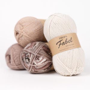 Könnte beinhalten: Vier Garnknäuel, die zusammen gestapelt sind. Ein cremefarbenes Knäuel im Vordergrund trägt ein braunes Papieretikett mit dem Text DROPS Fabel UNI COLOUR Superwash treated sock wool. Die anderen drei Knäuel sind in Beige-, Sand- und Brauntönen.