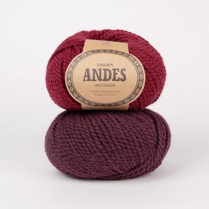Puede incluir: Dos ovillos de lana gruesa apilados en tonos rojo intenso y morado oscuro. El ovillo superior tiene una etiqueta de papel color canela con el texto DROPS ANDES UNI COLOUR, A soft and chunky blend of alpaca and wool, impreso en tinta negra.
