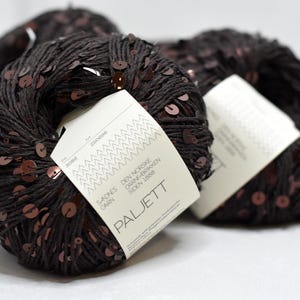 May include: Three balls of dark brown yarn embellished with small, reflective copper-colored sequins. Each ball features a white paper label with the text Sandnes Garn, Den Norske Garnfabrikken Siden 1888, and Paljett printed in black ink.