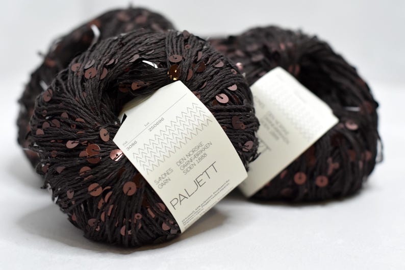 May include: Three balls of dark brown yarn embellished with small, reflective copper-colored sequins. Each ball features a white paper label with the text Sandnes Garn, Den Norske Garnfabrikken Siden 1888, and Paljett printed in black ink.