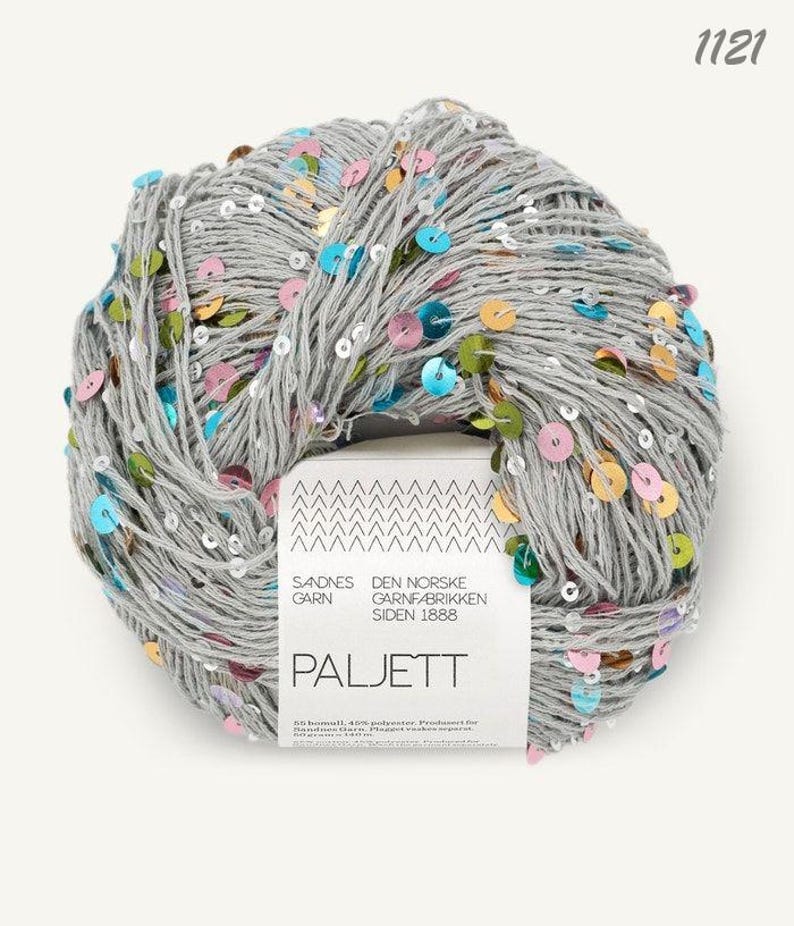 May include: A ball of light gray yarn interwoven with small, round, multicolored sequins in shades of blue, pink, green, and gold. A white paper label wraps around the center, featuring the text 1121, SANDNES GARN, and PALJETT.