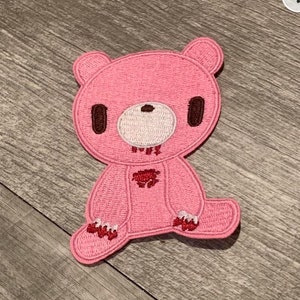 Peut inclure: Patch brodé rose d'un ours avec des yeux bruns, un nez blanc et du sang rouge qui coule. L'ours est assis les jambes croisées.