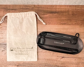 May include: A black metal tray with two black metal tongs. The tongs are labelled "Just Breathe Aroma" and the tray has the same logo. The tray is sitting on a wooden surface next to a white drawstring bag with the same logo.