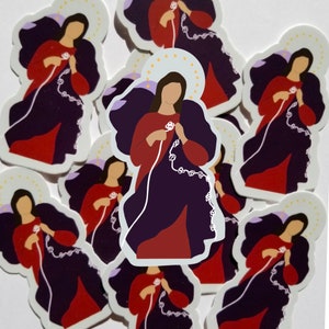 May include: A collection of stickers featuring a stylized illustration of a figure with a halo, wearing a red top and a dark purple cloak. The figure is holding a white rosary. The stickers have a white border and are scattered on a white surface.