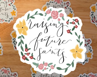 May include: White sticker with the text "raising future saints" in elegant black script, surrounded by a floral wreath of pink roses, yellow flowers, and green leaves. Several similar stickers are scattered on a wooden surface.