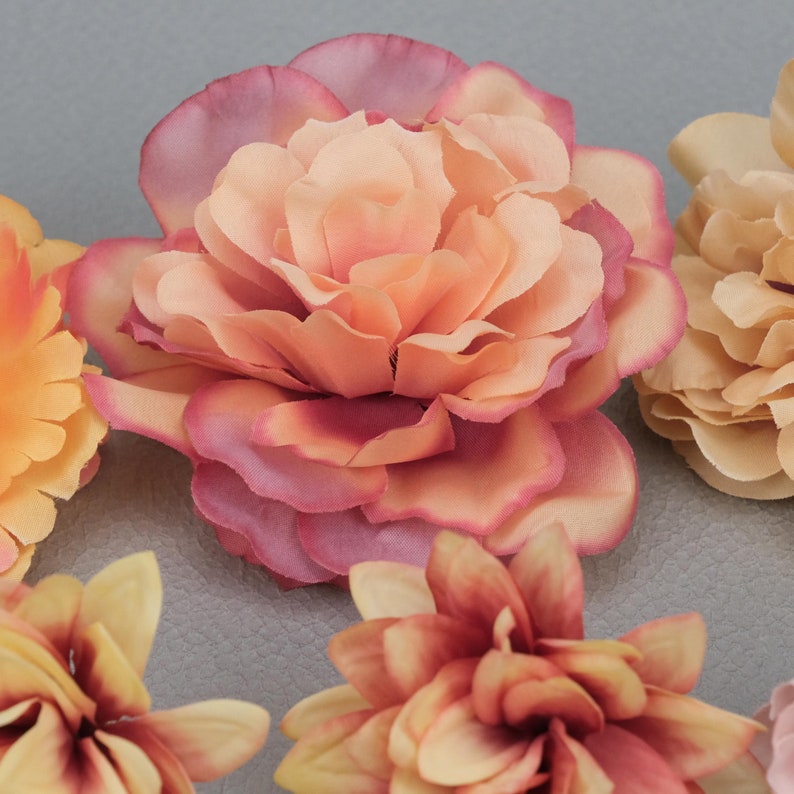 May include: Close-up of several artificial flowers in shades of peach, pink, and yellow. The central flower is a large, multi-layered bloom with a gradient of colors. Other flowers are visible, showcasing different petal arrangements.