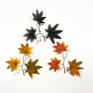 Puede incluir: Ramas de hojas de arce artificiales en negro, amarillo, naranja y verde. Las hojas tienen vetas detalladas y están dispuestas sobre finos tallos marrones. Las hojas están dispuestas en un patrón circular sobre un fondo blanco.