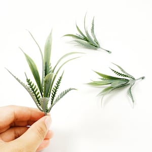 Puede incluir: Tres tallos de plantas verdes artificiales con diferentes formas de hojas se muestran sobre un fondo blanco. Un tallo es sostenido por una mano. Las hojas varían en color de verde claro a oscuro, con texturas detalladas.