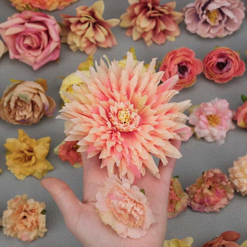 May include: A collection of artificial flowers in various shades of pink, peach, and yellow. The image features a large, multi-petaled peach-colored flower held in a hand, with other flowers scattered around. The flowers appear to be made of fabric and are likely for crafting or decoration.