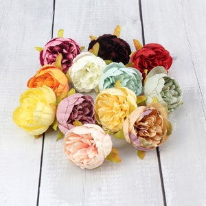 May include: A collection of 12 artificial peony flower heads in various colors, including pink, white, yellow, orange, red, and burgundy. The flowers are arranged in a cluster on a white wooden surface.