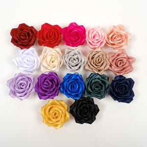 Op de afbeelding: Een verzameling kleurrijke satijnen lintrozen in verschillende tinten, waaronder rood, roze, paars, blauw en goud. De rozen zijn in rijen gerangschikt op een witte ondergrond, waarbij hun gedetailleerde bloemblaadjesontwerp wordt getoond. Ideaal voor knutselen en versieringen.