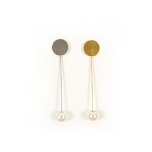 May include: A pair of dangle earrings. One earring features a silver-colored disc, a thin silver rod, and a white pearl. The other earring has a gold-colored disc, a thin gold rod, and a white pearl. The earrings are displayed on a white background.