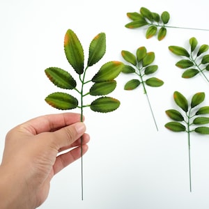Op de afbeelding: Kunstmatige groene bladeren met oranje getipte uiteinden op dunne, donkergroene stelen. Eén steel wordt door een hand vastgehouden, andere zijn verspreid op een witte achtergrond. Deze kunstbladeren zijn geschikt voor knutselen of decoratie. De stelen zijn ongeveer 15 cm lang.
