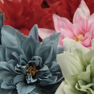 May include: A close-up of three artificial flowers. The flowers are made of fabric and have a realistic appearance. The flower on the left is a blue dahlia, the flower in the centre is a pink dahlia, and the flower on the right is a green dahlia.