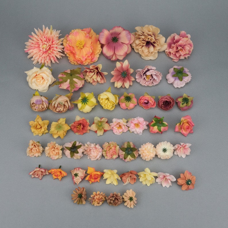 May include: Assortment of artificial flowers in various colors, including pink, peach, yellow, and orange. The flowers are arranged on a gray surface, showcasing different shapes and sizes, ideal for crafting or decorative projects.