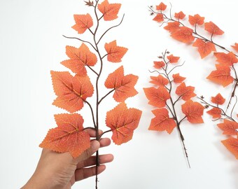 May include: A bunch of artificial autumn leaves with a brown stem and orange leaves. The leaves are arranged in a cluster and are perfect for decorating for the autumn season.