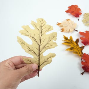 May include: A single, light brown, artificial leaf with a detailed vein pattern. The leaf is held in a hand against a white background.