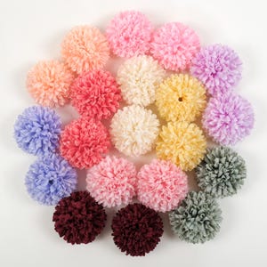 May include: A collection of colorful, round artificial flowers in various shades of pink, peach, yellow, purple, blue, cream, burgundy, and green. The flowers are arranged in a circular pattern against a white background.