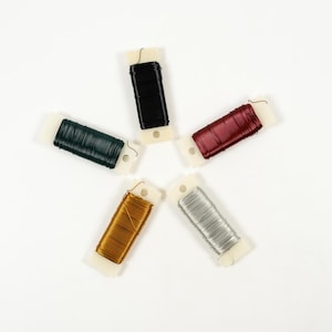 Puede incluir: Vista aérea de cinco carretes de alambre floral dispuestos en forma de estrella. Los colores del alambre incluyen negro, verde oscuro, burdeos, dorado y plateado. Cada carrete está montado sobre una base de plástico blanco con un pequeño agujero.