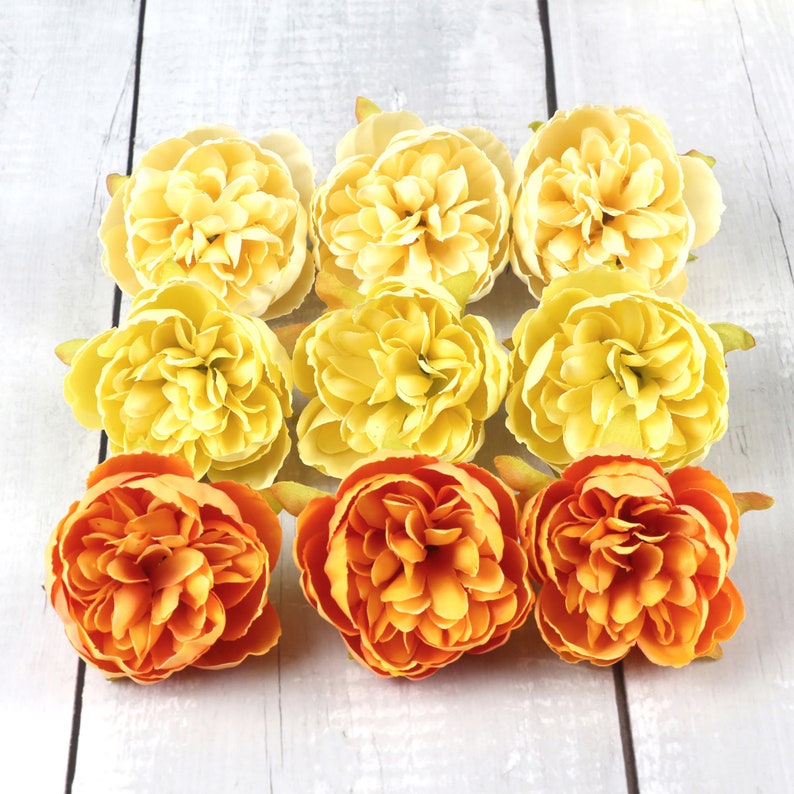 May include: Nine artificial flower heads in two colors: six yellow and three orange. The flowers are arranged in a circle on a white wooden surface.
