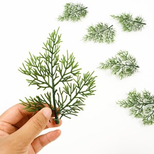 May include: Close-up of several artificial green fern stems. One stem is held by a hand, while others are scattered on a white surface. The stems have a detailed, realistic appearance, with many small, delicate leaves.