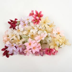 May include: A collection of colorful artificial flowers in shades of pink, red, yellow, and white. The flowers are arranged in a loose cluster, with water droplets visible on the petals. The flowers are on a white background.