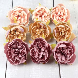 May include: Nine artificial flowers in three different colors: peach, light brown, and mauve. The flowers are arranged in a circle on a white wooden surface.
