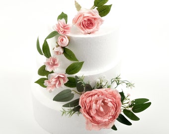 Op de afbeelding: Een drielaagse witte taartdummy versierd met roze en groene bloemenaccenten. De taart is versierd met een waterval van roze rozen en kleinere roze bloemen, afgewisseld met groene bladeren.