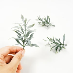 Op de afbeelding: Verschillende kunstmatige plantenstelen met groene en grijze bladeren worden getoond tegen een witte achtergrond. Eén stengel wordt door een hand vastgehouden. De bladeren hebben een textuur en de stelen zijn slank.