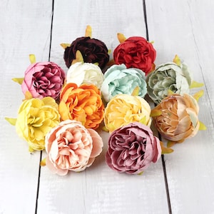 May include: Assortment of artificial silk peonies in various colors, including pink, peach, yellow, orange, red, burgundy, and green. The flowers are arranged in a cluster on a white wooden surface.