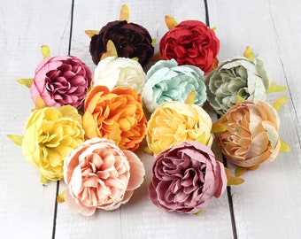 May include: Assortment of artificial silk peonies in various colors, including pink, peach, yellow, orange, red, burgundy, and green. The flowers are arranged in a cluster on a white wooden surface.