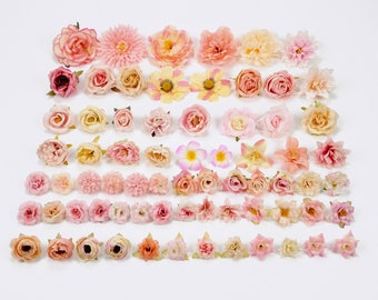 May include: An assortment of artificial flowers in various shades of pink, peach, and yellow, arranged on a white surface. The collection includes roses, daisies, and other floral designs, perfect for crafting or decoration.