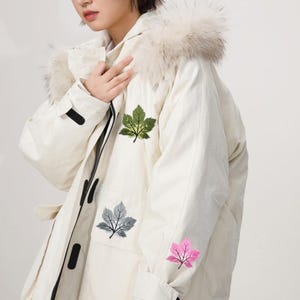 May include: Cream-colored winter coat with a fur-trimmed hood. The coat features three embroidered leaf designs in green, gray, and pink. The coat has a zipper closure and black accents.