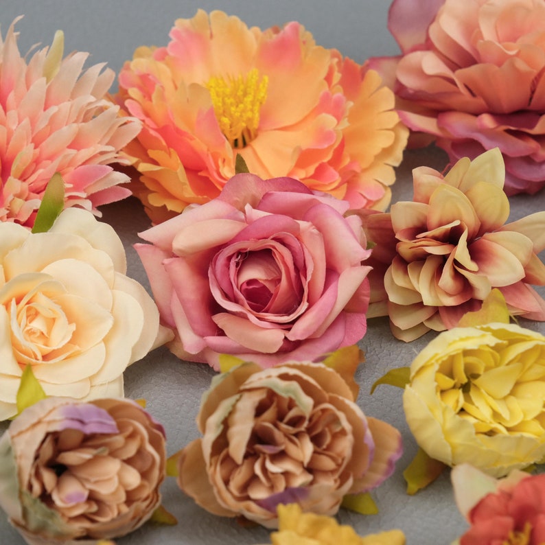 May include: A collection of colorful artificial flowers in shades of peach, pink, and yellow. The arrangement includes roses, peonies, and other floral varieties, with green stems and leaves. The flowers are arranged on a gray surface.