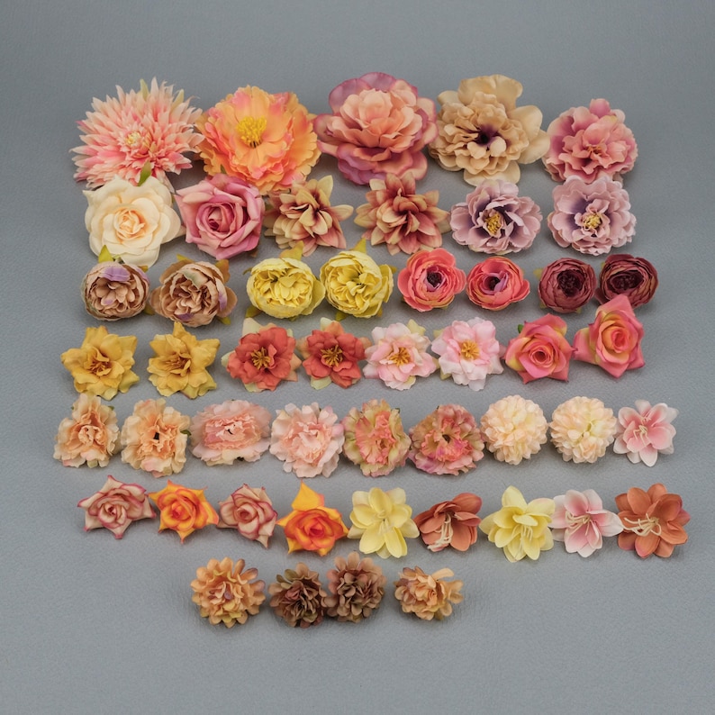 May include: A collection of artificial flowers in various sizes and colors, including pink, peach, yellow, and orange. The assortment features different flower types, such as roses and peonies, arranged on a gray surface.