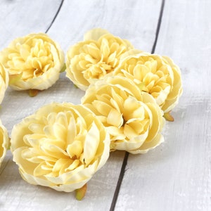 May include: A group of six pale yellow artificial flowers with ruffled petals. The flowers are arranged in a circle on a white wooden surface.