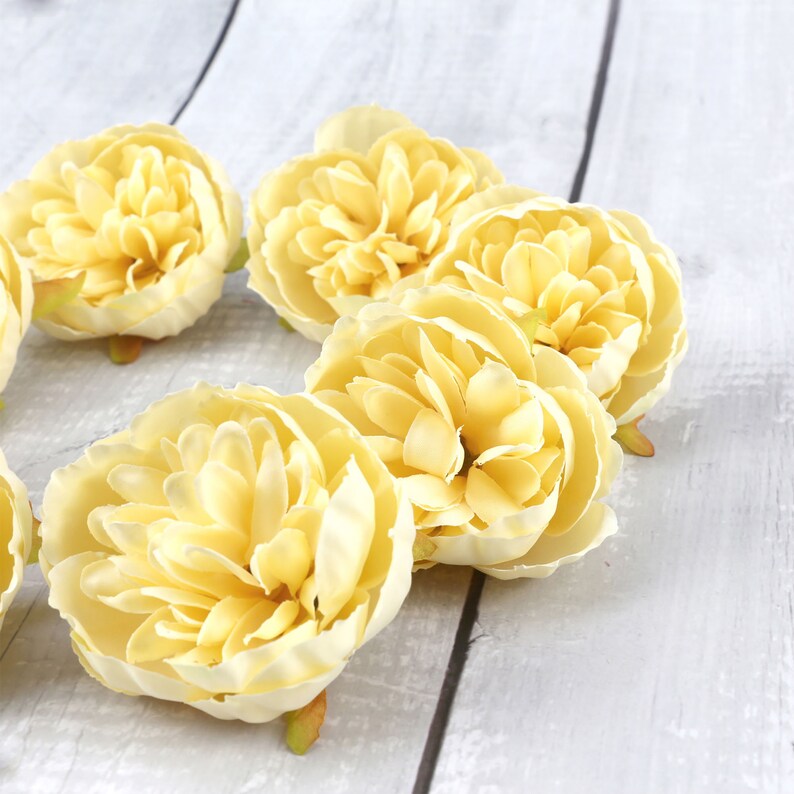 May include: A group of six pale yellow artificial flowers with ruffled petals. The flowers are arranged in a circle on a white wooden surface.
