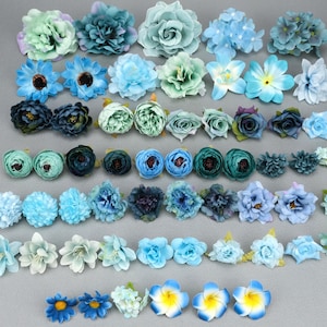 May include: A collection of artificial flowers in various shades of blue and green. The assortment includes roses, daisies, and other floral designs, perfect for crafting or decorative purposes. The flowers are arranged on a neutral gray surface.