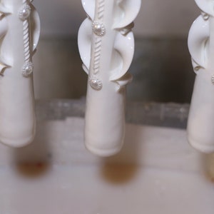 Pu&ograve; includere: Tre candele bianche ornate con dettagli di perle vengono immerse in un liquido bianco. Le candele sono appese a un supporto metallico.
