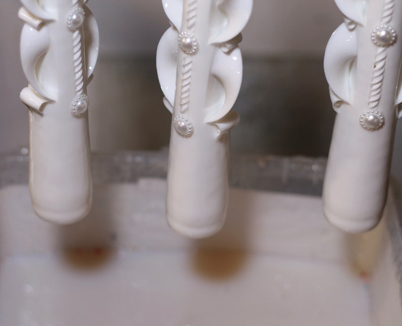 Pu&ograve; includere: Tre candele bianche ornate con dettagli di perle vengono immerse in un liquido bianco. Le candele sono appese a un supporto metallico.