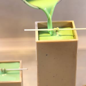 May include: Green wax is being poured into a square candle mold with a wick centered in the mold. The mold is made of a light beige material.