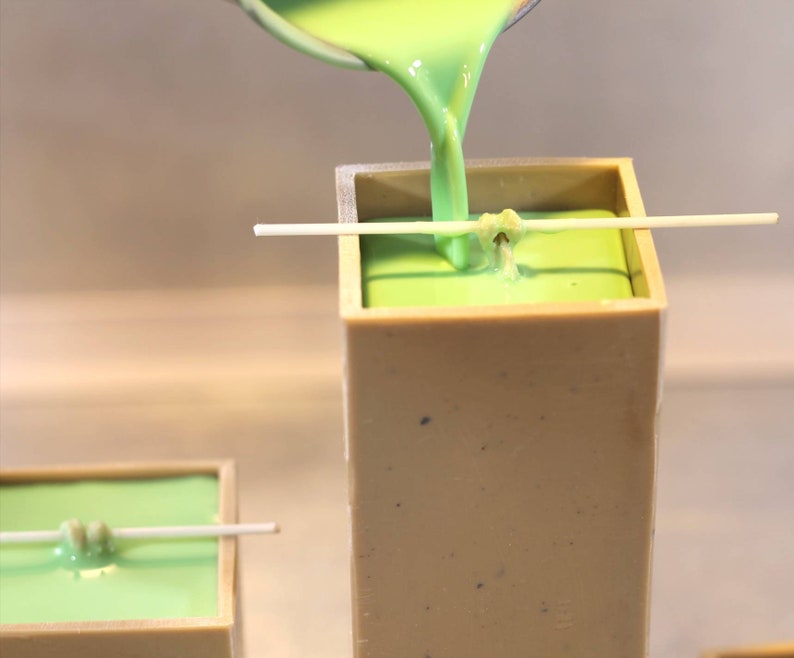 May include: Green wax is being poured into a square candle mold with a wick centered in the mold. The mold is made of a light beige material.