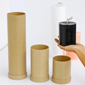 May include: Three beige cylindrical containers with a speckled texture, and a black cylindrical candle with a coiled wick. The containers are arranged in a row, with the candle in front of the middle container.