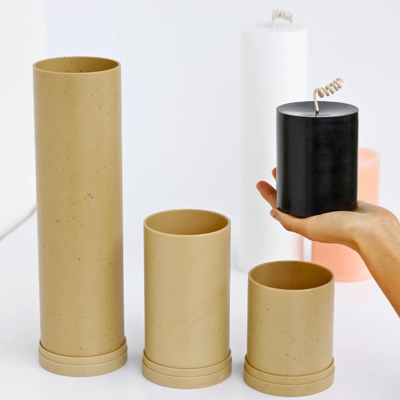 May include: Three beige cylindrical containers with a speckled texture, and a black cylindrical candle with a coiled wick. The containers are arranged in a row, with the candle in front of the middle container.
