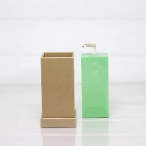 May include: A square candle mold made of beige silicone with a green pillar candle inside. The candle has a wick sticking out of the top.