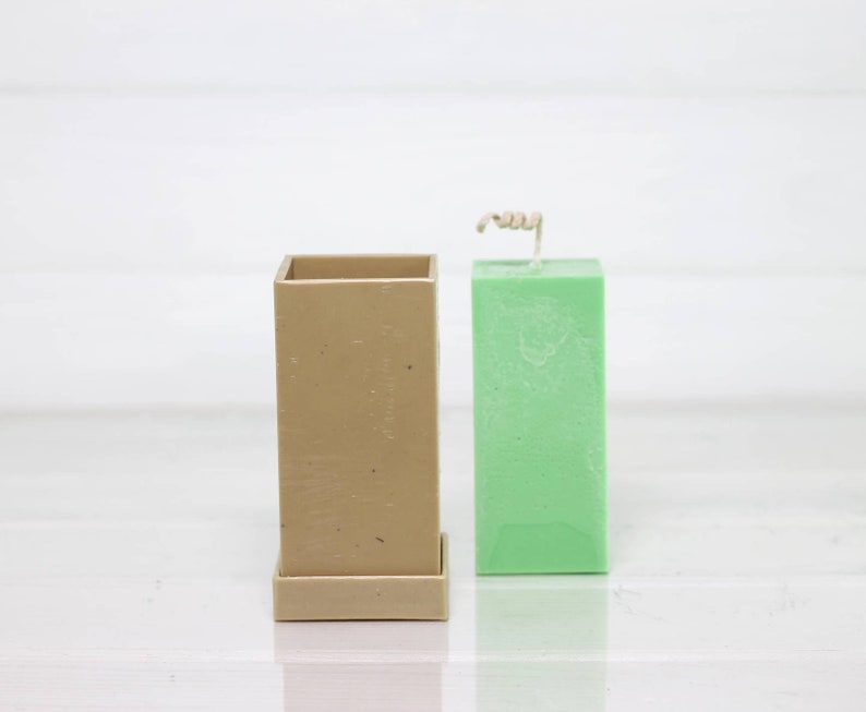 May include: A square candle mold made of beige silicone with a green pillar candle inside. The candle has a wick sticking out of the top.