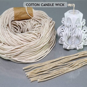 May include: A close-up of a spool of natural cotton candle wicks. The wicks are white and are wound around a brown paper core. There is a single wick laid out straight on the table next to the spool. The text "COTTON CANDLE WICK" is printed in black on a white background at the top of the image.