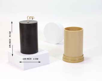 Puede incluir: Una vela negra en forma de pilar con una mecha, que mide 10 cm de alto y 6 cm de ancho, se muestra sobre una base cuadrada blanca. Un molde de vela cilíndrico beige con un diámetro de 6 cm y una altura de 10 cm se muestra junto a la vela.